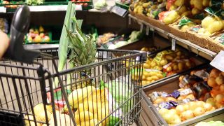 Ein Einkaufswagen ist bodenbedeckt mit Gemüde und Obst. Im Hintergrund ist die Obst- und Gemüseabteilung eines Supermarktes zu sehen. | Quelle: IMAGO / CHROMORANGE